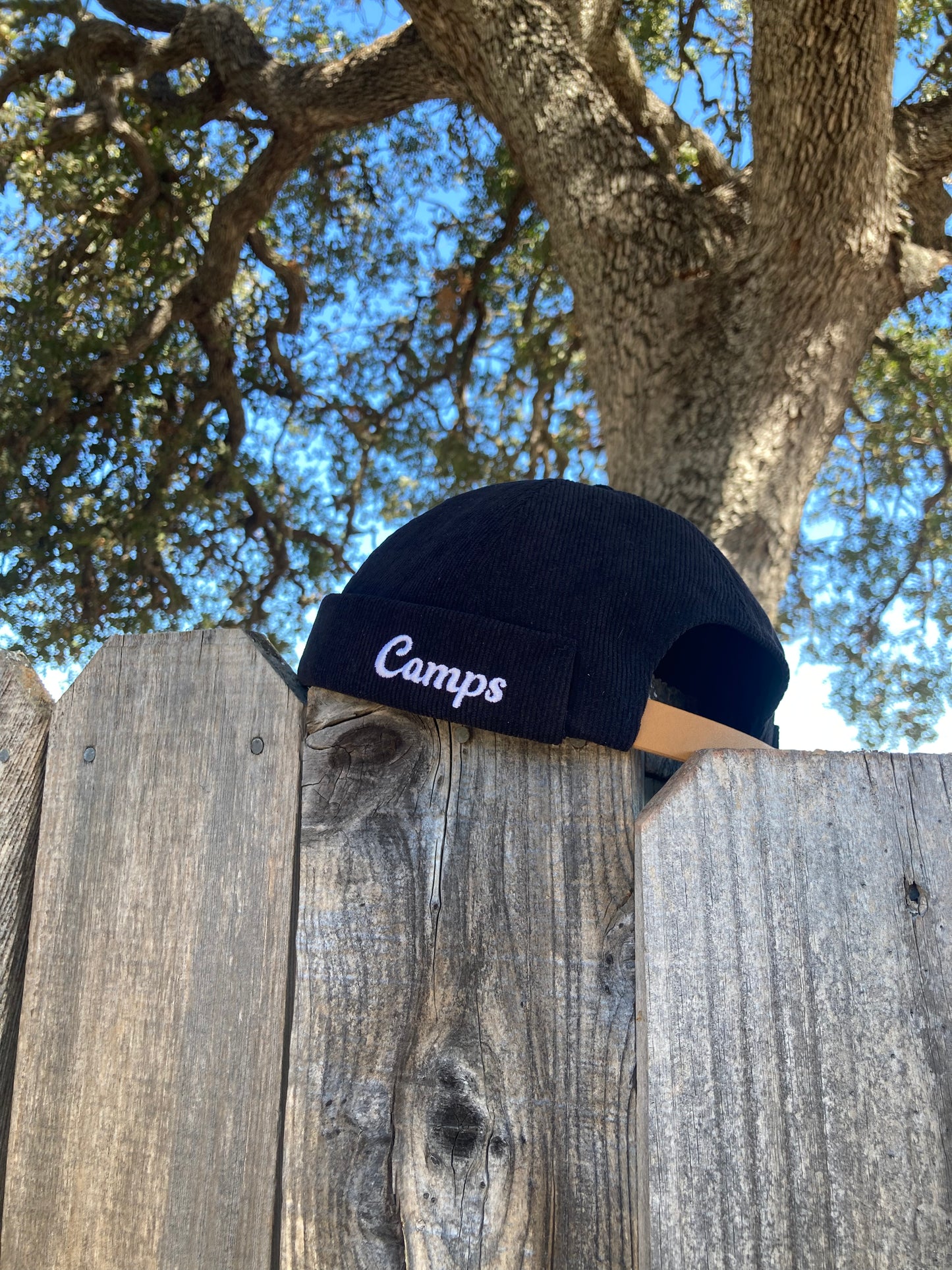 Black cap with 'Camps' text on a wooden fence with trees in the background