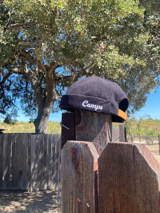 Black cap with 'Camps' logo on a wooden post outdoors