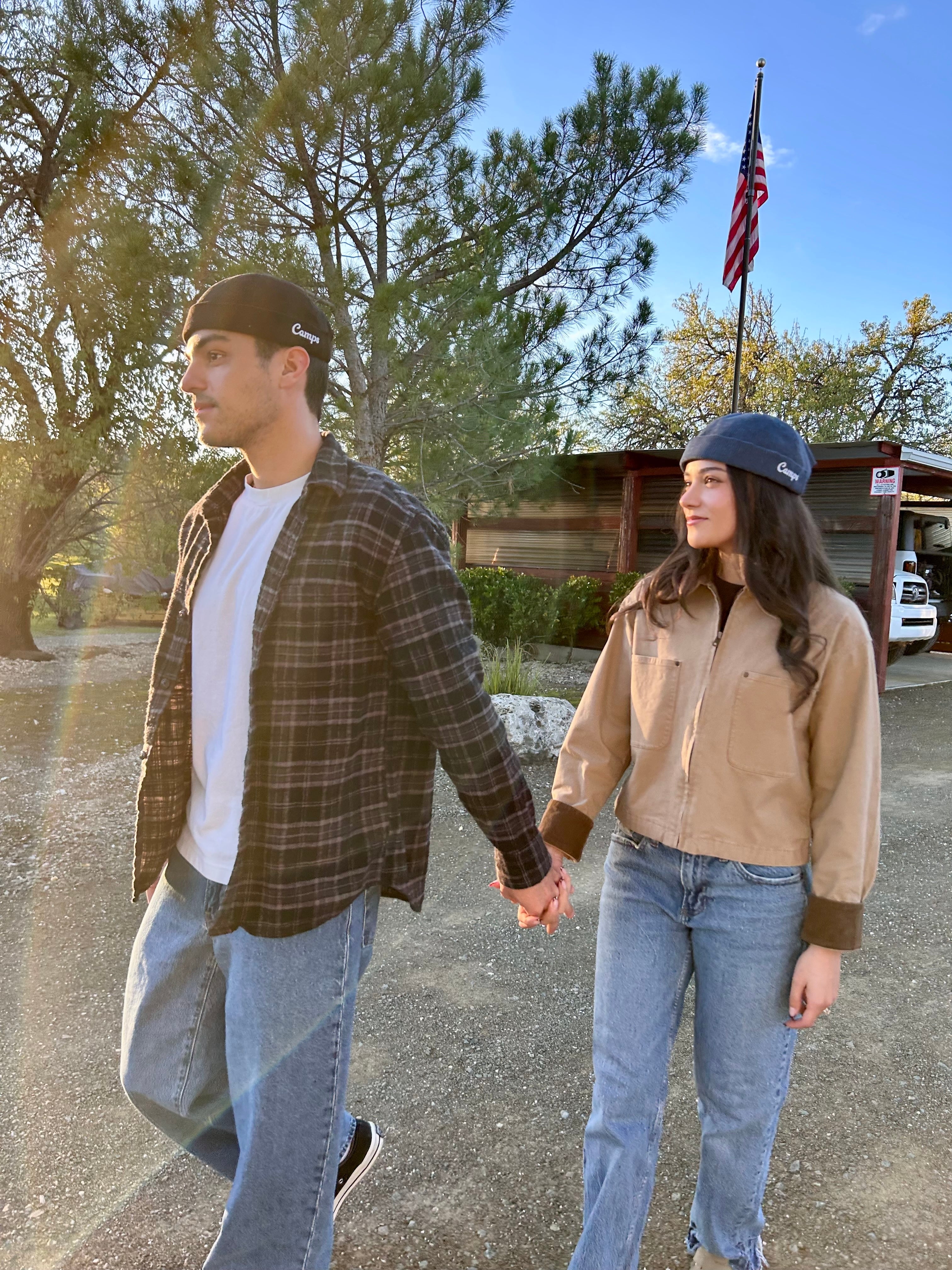 Two people holding hands on a road with trees and a building in the background wearing Camps corduroy brimless hats.