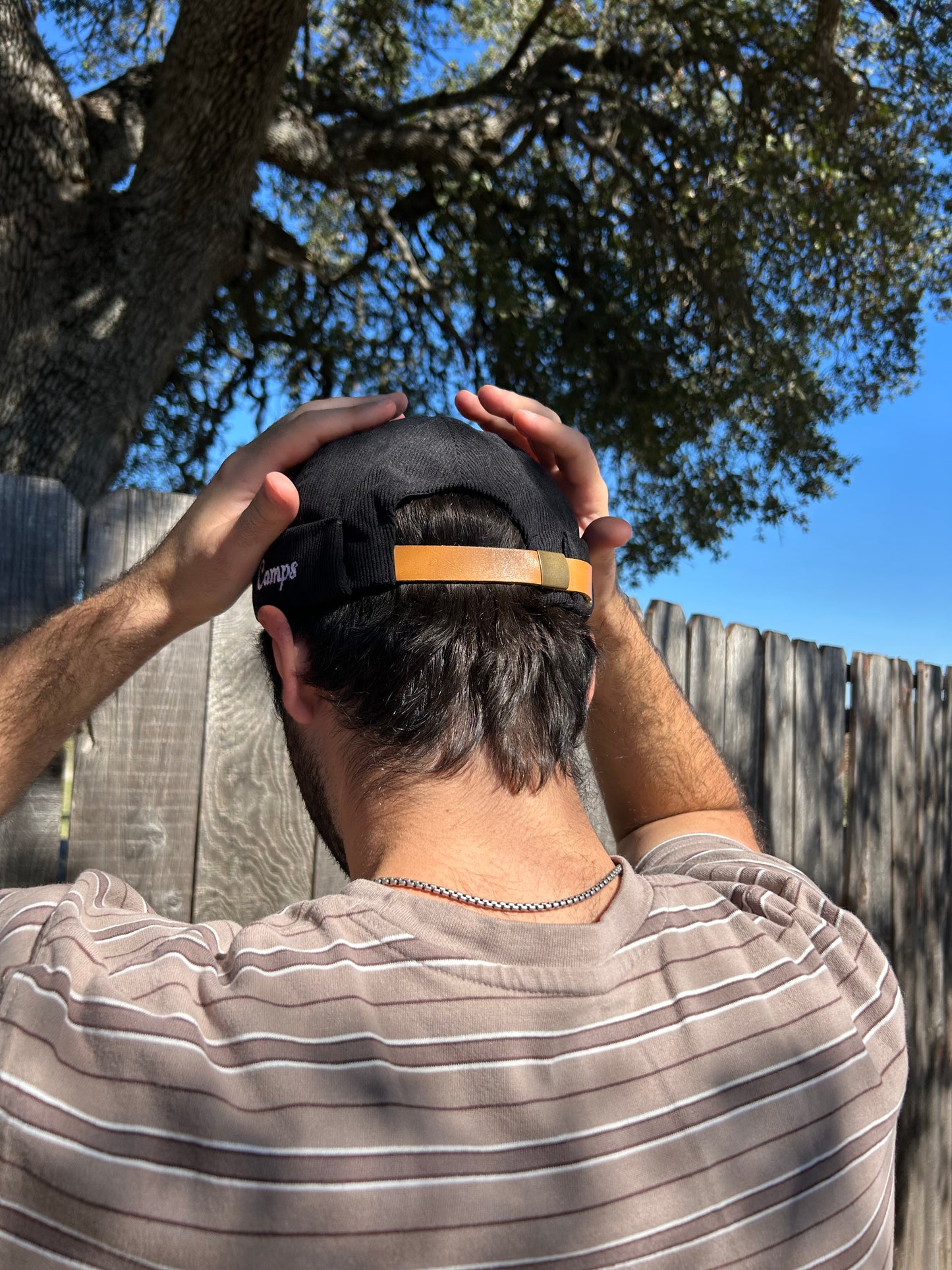 Person adjusting a brimless cap outdoors with trees and a fence in the background