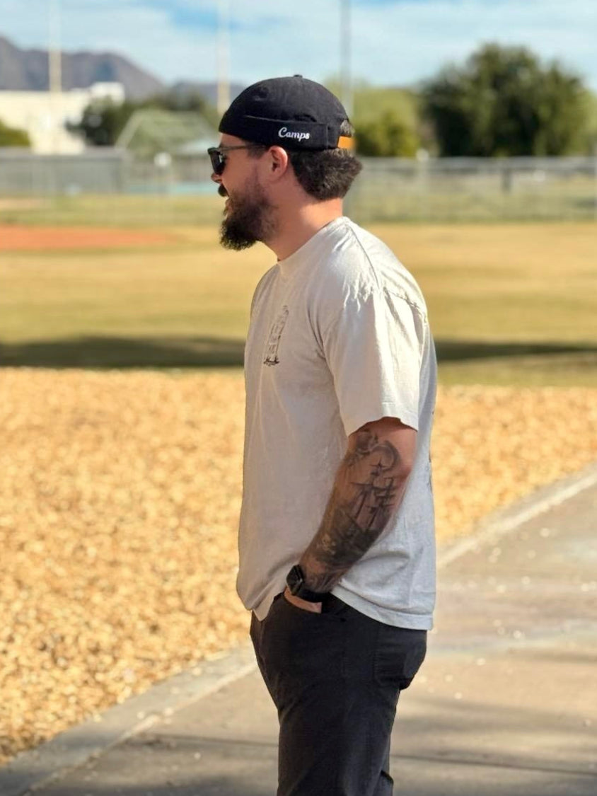 Man with tattoos and a beard wearing sunglasses and a brimless cap, standing on a path with grass and trees in the background.