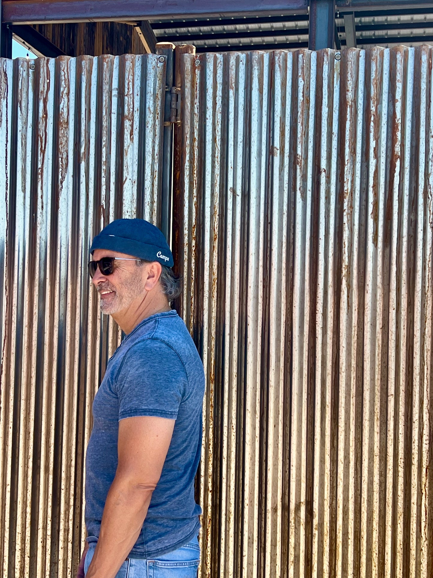 Man wearing a blue brimless hat cap and sunglasses leaning against a corrugated metal wall.