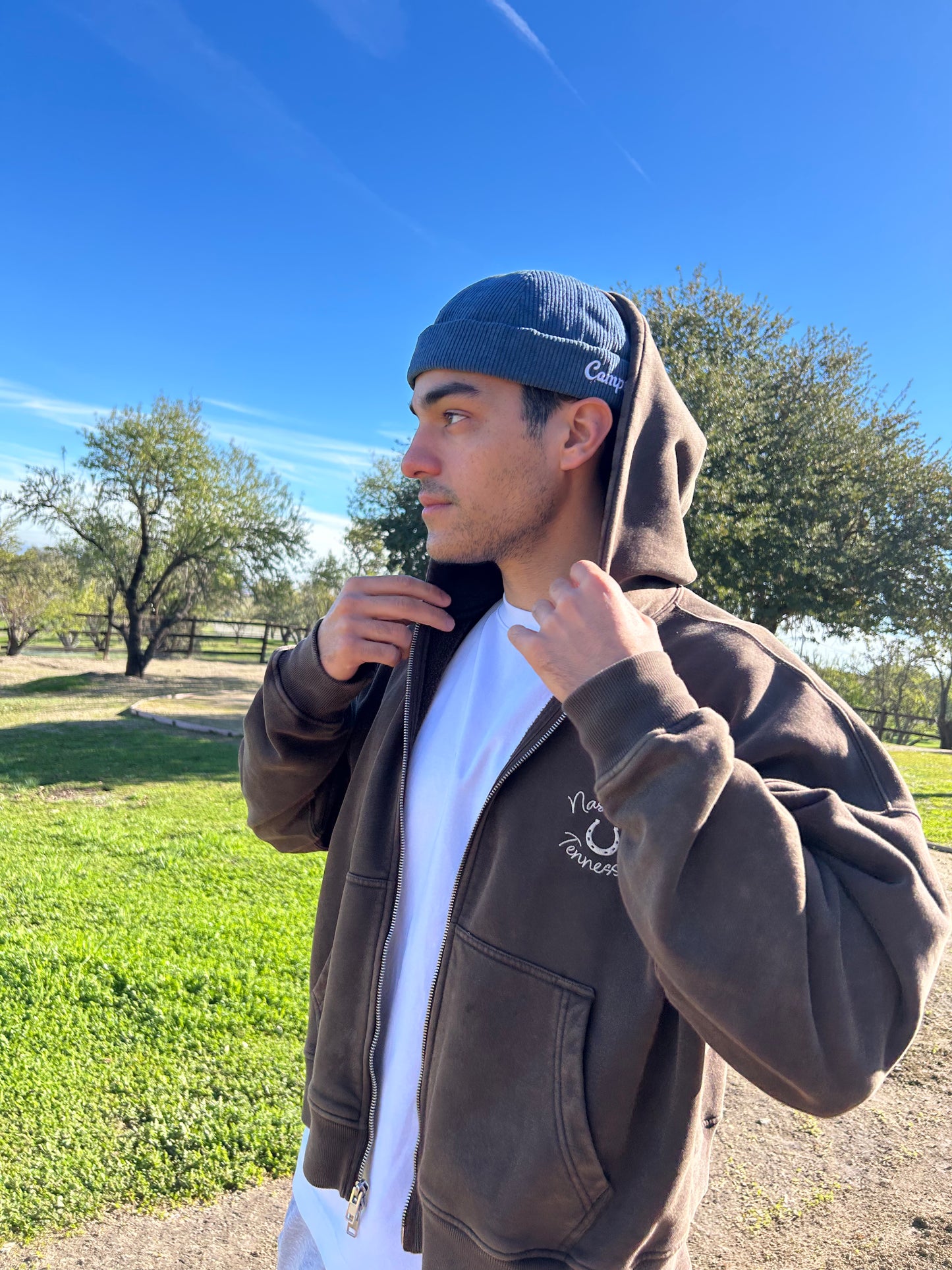Man wearing a brown hoodie and navy brimless cap outdoors with trees and clear sky in the background