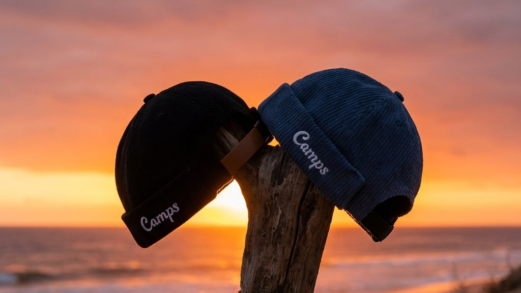 Two brimless hats / docker caps with 'Camps' branding on a wooden stick against a sunset ocean backdrop.